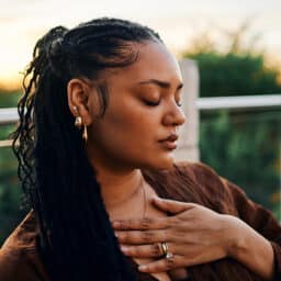 A serene woman with braided hair and golden accessories sits outdoors at sunset, her hand over her chest, exuding calm and introspection.