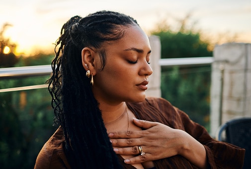 A serene woman with braided hair and golden accessories sits outdoors at sunset, her hand over her chest, exuding calm and introspection.
