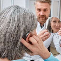 Senior woman trying on a hearing aid while looking into a mirror held by a hearing aid specialist in audiology clinic.