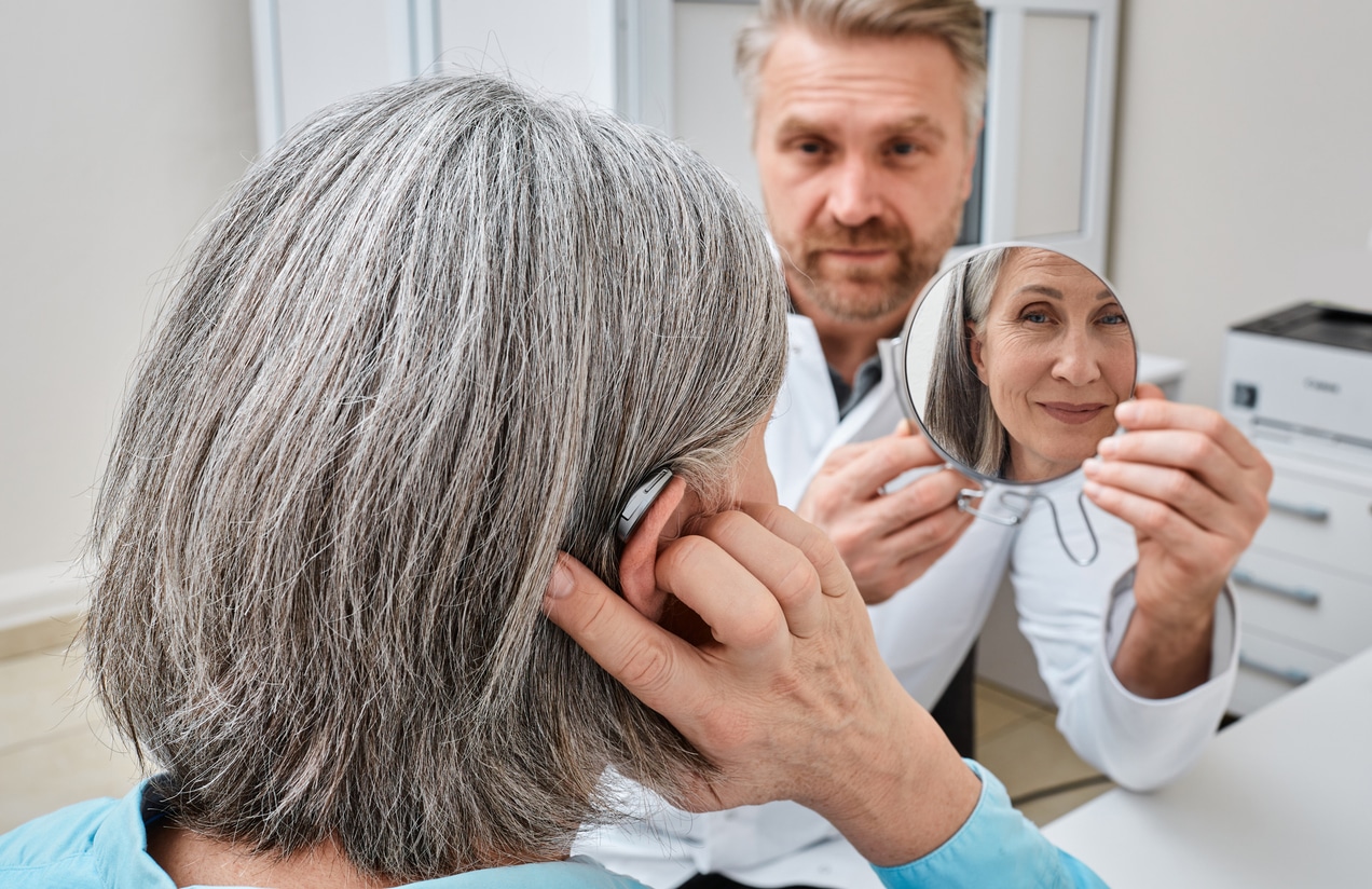 Senior woman trying on a hearing aid while looking into a mirror held by a hearing aid specialist in audiology clinic.