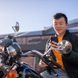 A man sits on a parked motorcycle while looking at his smartphone in an parking lot.
