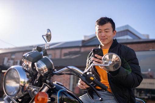 A man sits on a parked motorcycle while looking at his smartphone in an parking lot.